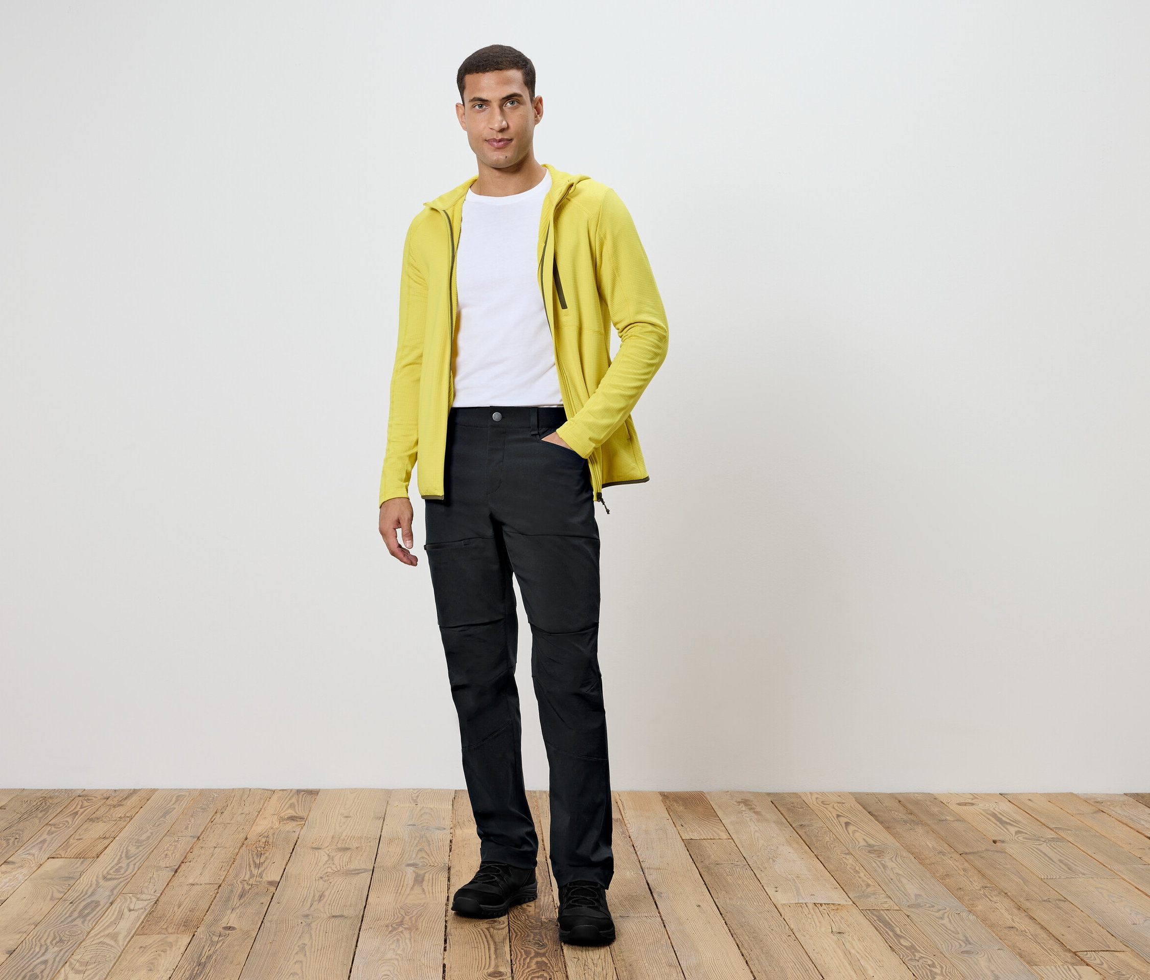 Mann steht auf Holzboden. Er trägt eine schwarze Hose, ein weißes T-Shirt und eine gelbe Kapuzenjacke.