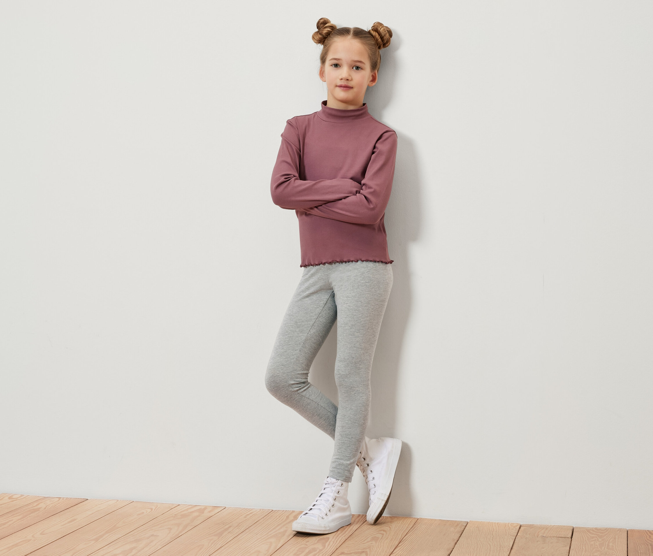 Ein Mädchen mit braunen, zu Duttfrisuren hochgesteckten Haaren steht mit verschränkten Armen vor einer Wand. Sie trägt ein weinrotes Langarmshirt, graue Leggings und weiße Turnschuhe.