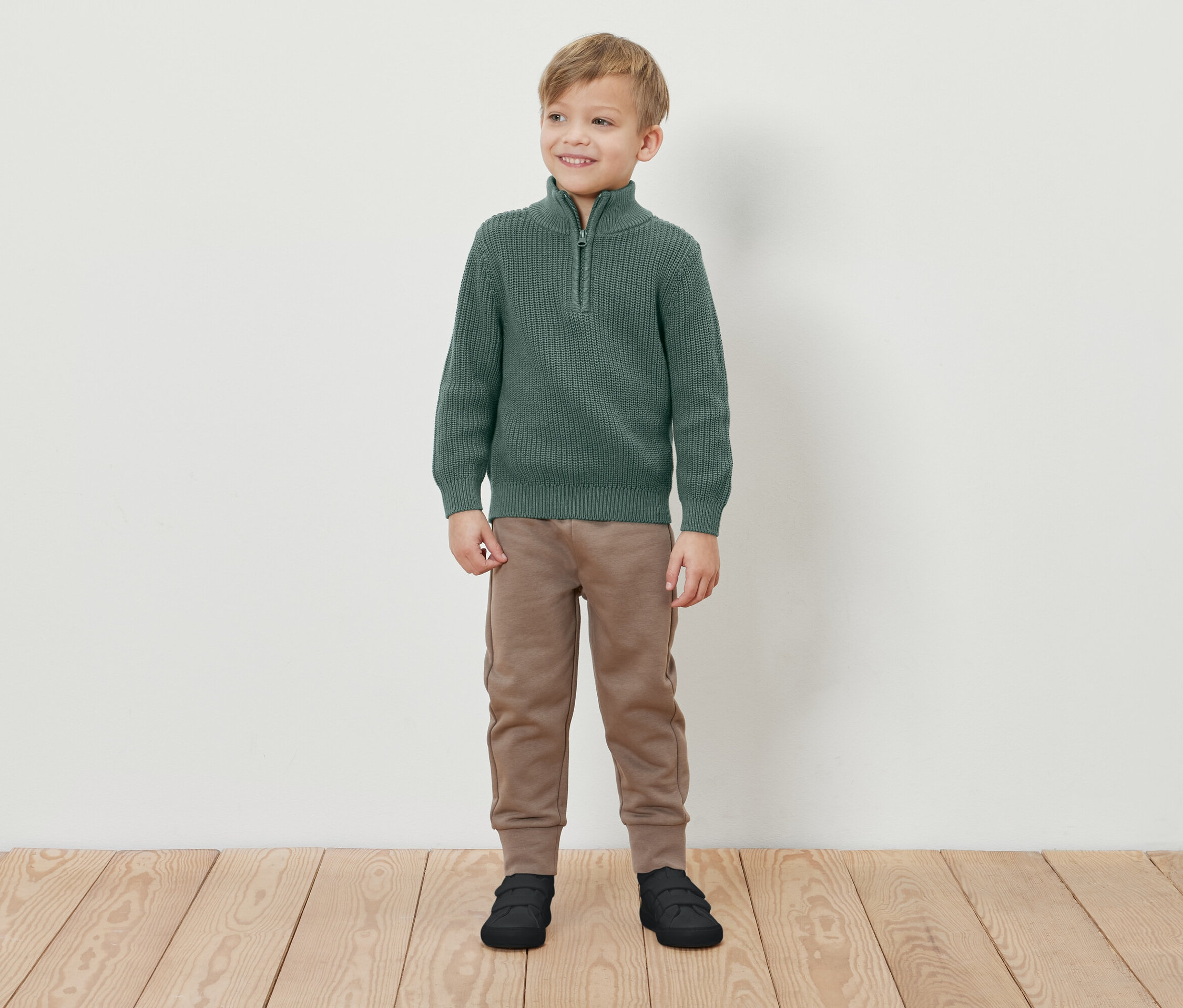 Ein Junge steht lächelnd auf einem Holzboden und trägt einen grünen Pullover mit Reißverschluss, braune Jogginghose und schwarze Schuhe.