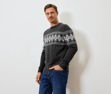 Mann posiert in grauem Pullover mit Muster und blauen Jeans.