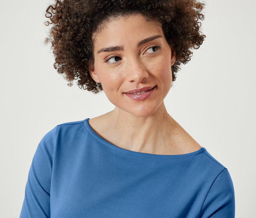 Frau mit braunen, lockigen Haaren trägt ein blaues Halbarmshirt.