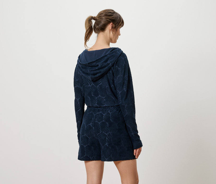 Rückansicht einer Frau mit hochgesteckten braunen Haaren, die einen marineblauen, gemusterten Kapuzenpullover und Shorts trägt.