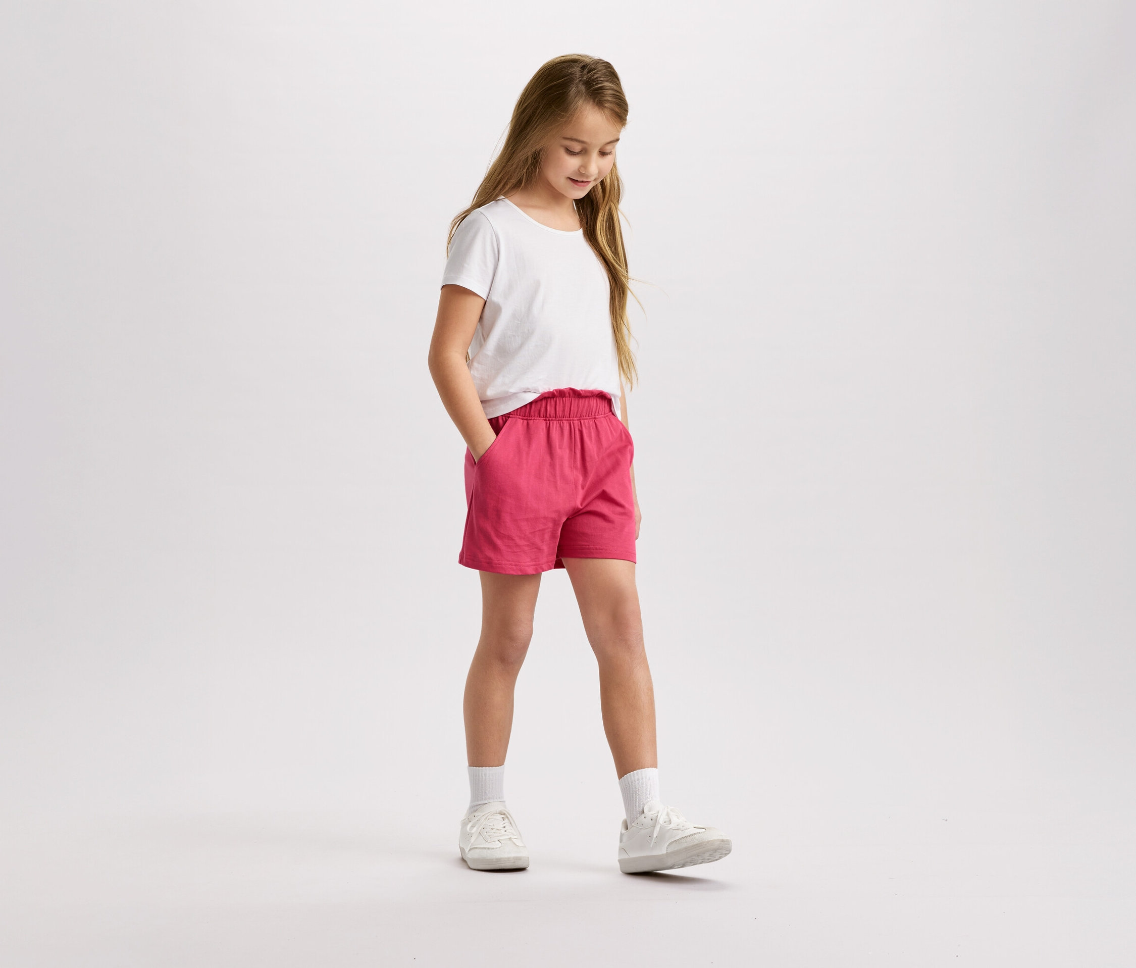 Mädchen mit blonden Haaren, weißem T-Shirt und pinkfarbenen Shorts steht da und schaut nach unten.