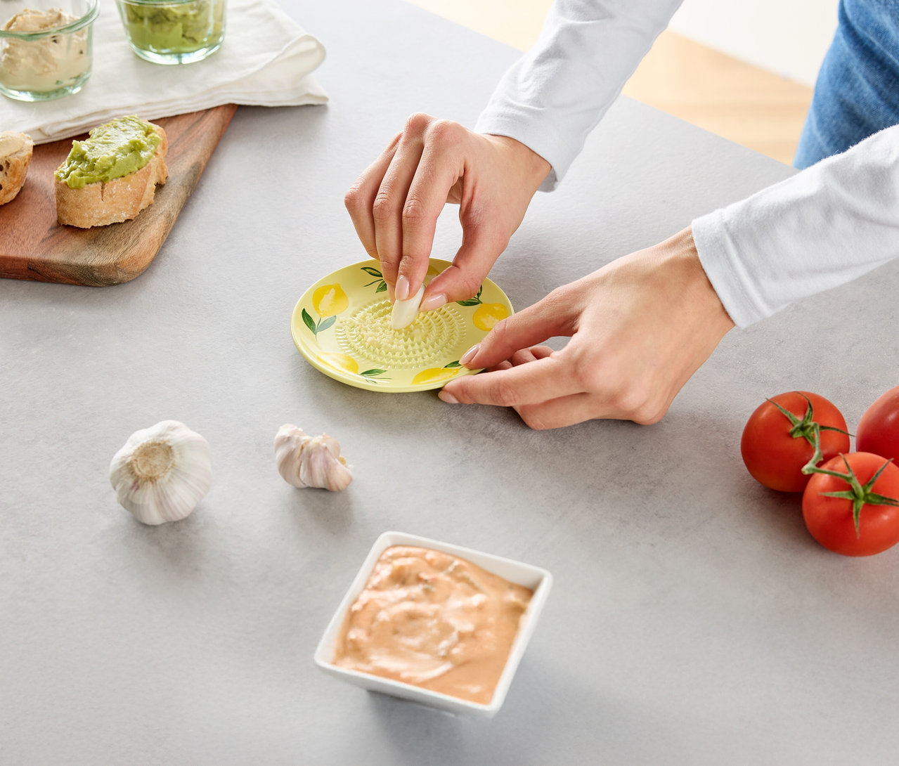 Eine Person reibt Knoblauch auf einer gelben Reibe, umgeben von Tomaten und Dips.