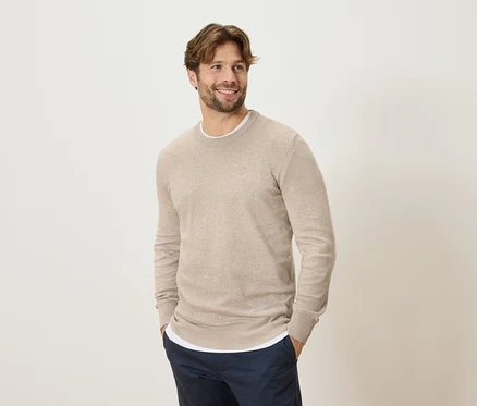 Mann mit beigem Pullover und blauen Hosen, die Hände in den Hosentaschen, lächelnd und seitlich blickend.