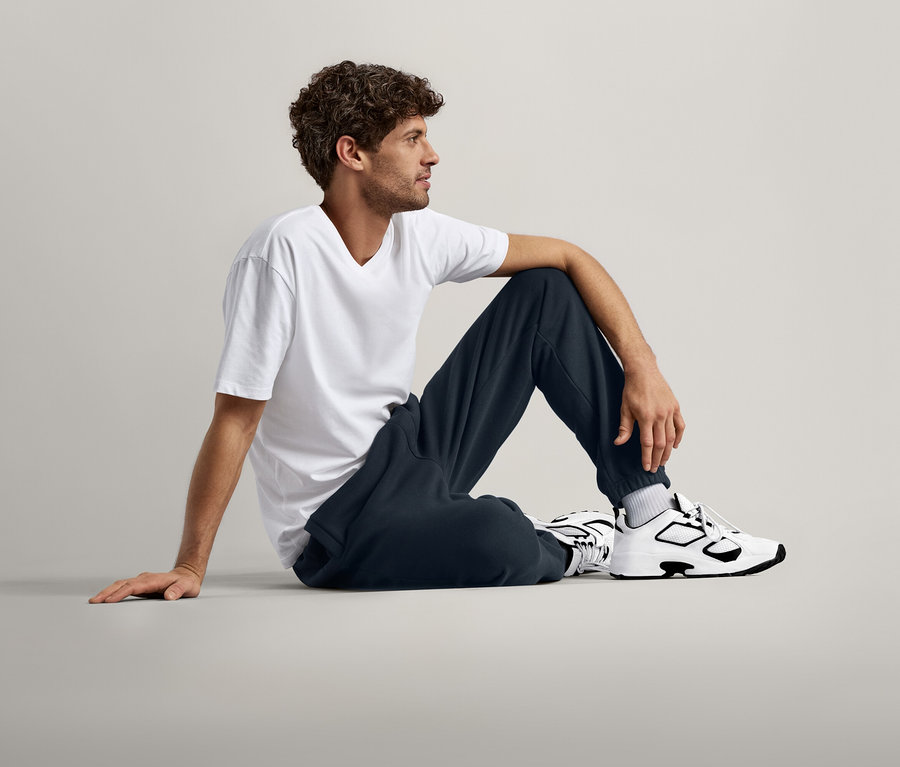 Mann mit lockigem Haar sitzt auf dem Boden, trägt ein weißes T-Shirt, dunkelblaue Jogginghose und weiße und schwarze Turnschuhe.
