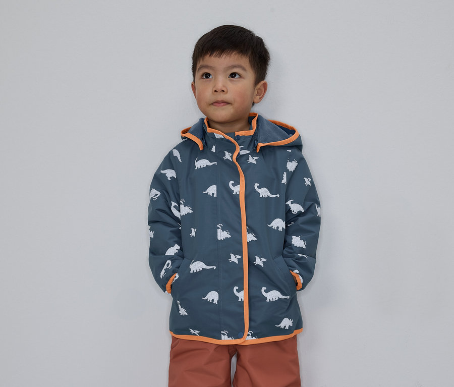 Ein Junge mit dunklen Haaren trägt eine blaue Jacke mit Dinosaurier-Muster und orangefarbenen Akzenten vor einem grauen Hintergrund.