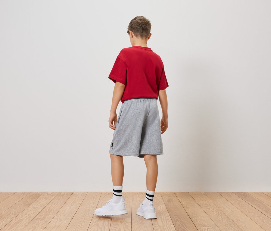Ein Jugendlicher steht mit dem Rücken zur Kamera, er trägt ein rotes T-Shirt, graue Shorts, weiße Socken und Turnschuhe.