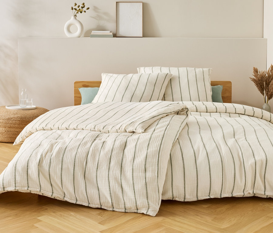 Bett mit gestreifter Bettwäsche, zwei Kissen und einer Decke. Auf dem Nachttisch stehen eine Vase und Gläser mit Wasser.