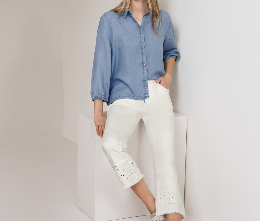 Frau steht, trägt eine hellblaue Street One Jeansbluse und eine weiße Street One Jeans mit Lochstickerei, lehnt an einer weißen Wand und einem Würfel.