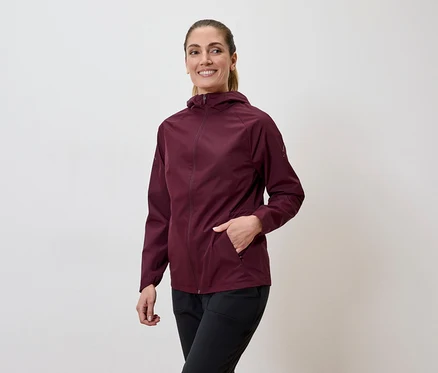 Frau mit Pferdeschwanz trägt weinrote Jacke mit Kapuze und schwarze Hose, Hand in der Jackentasche.