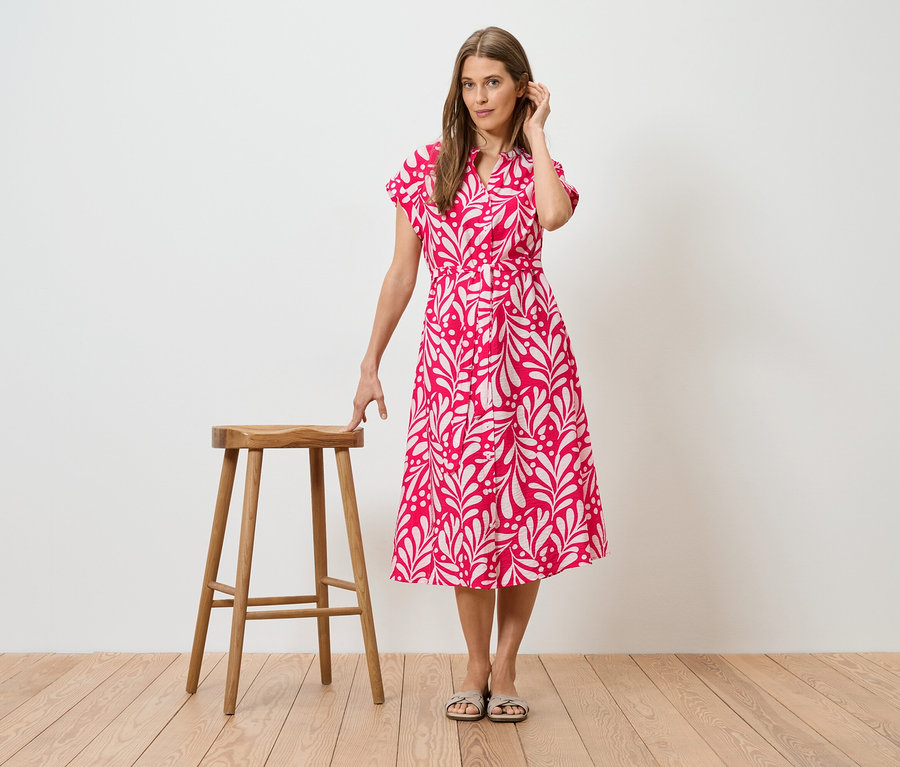 Frau im pinkfarbenen Kleid mit weißem Blattmuster, stehend neben einem Holzschemel.