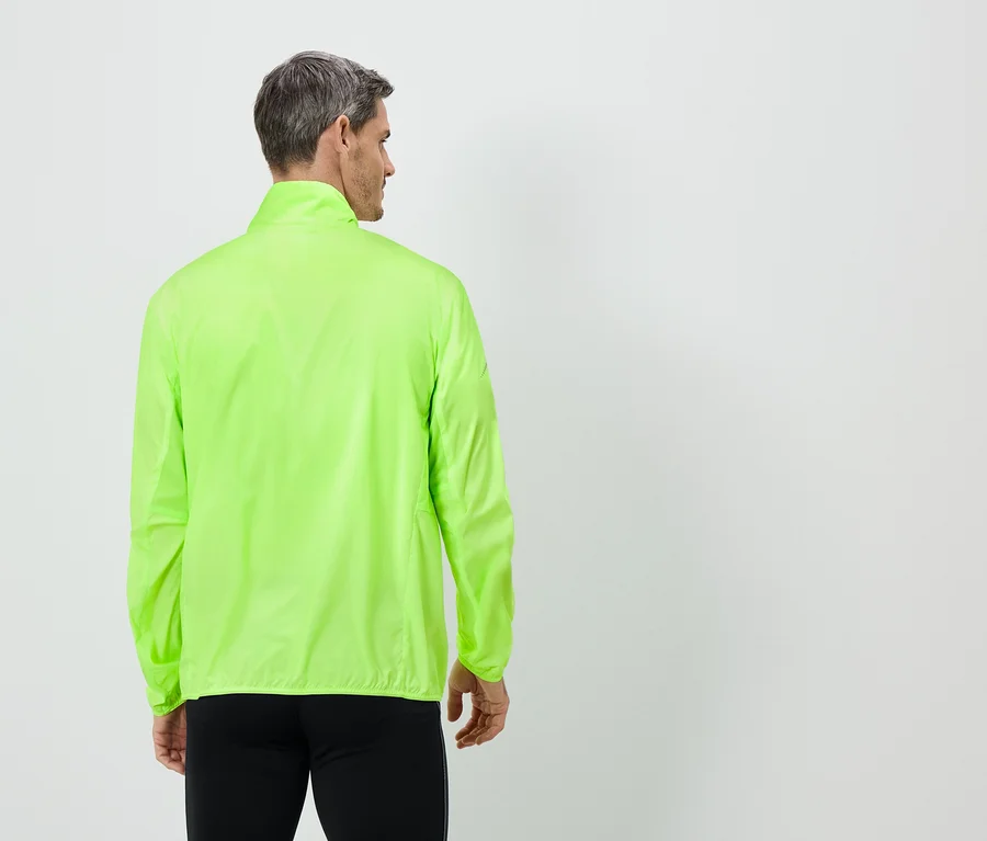 Ein Mann von hinten, mit grauen Haaren, trägt eine neongrüne Jacke und schwarze Hose vor einem weißen Hintergrund.