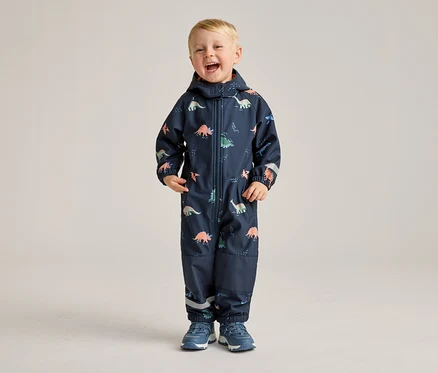 Ein lachender Junge steht in einem marineblauen Overall mit Dinosaurier-Muster und blauen Schuhen vor einem hellen Hintergrund.