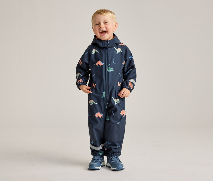 Ein lachender Junge steht in einem marineblauen Overall mit Dinosaurier-Muster und blauen Schuhen vor einem hellen Hintergrund.