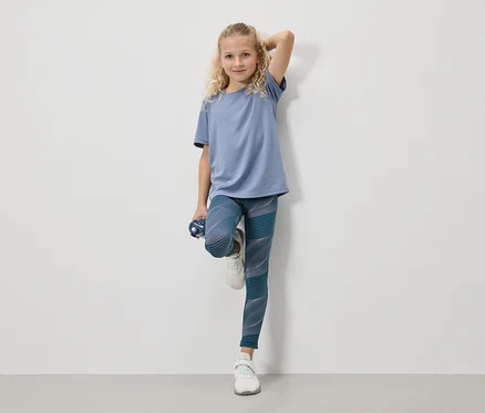 Ein blondes Mädchen in einem blauen T-Shirt, Leggings und weißen Turnschuhen steht mit einer Wasserflasche vor einer weißen Wand.