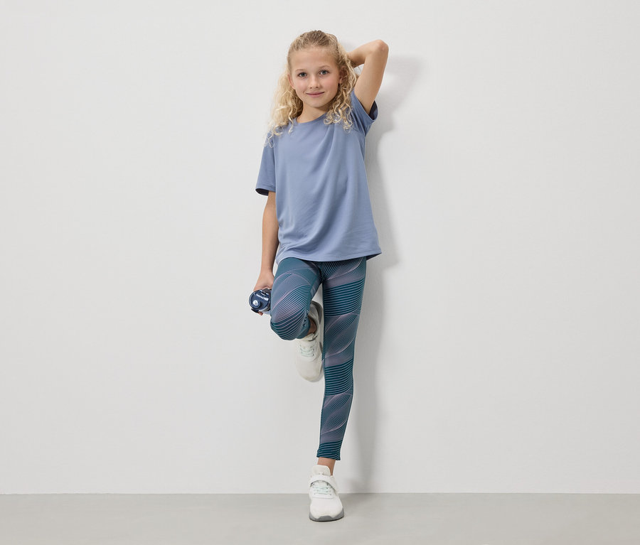 Ein blondes Mädchen in einem blauen T-Shirt, Leggings und weißen Turnschuhen steht mit einer Wasserflasche vor einer weißen Wand.