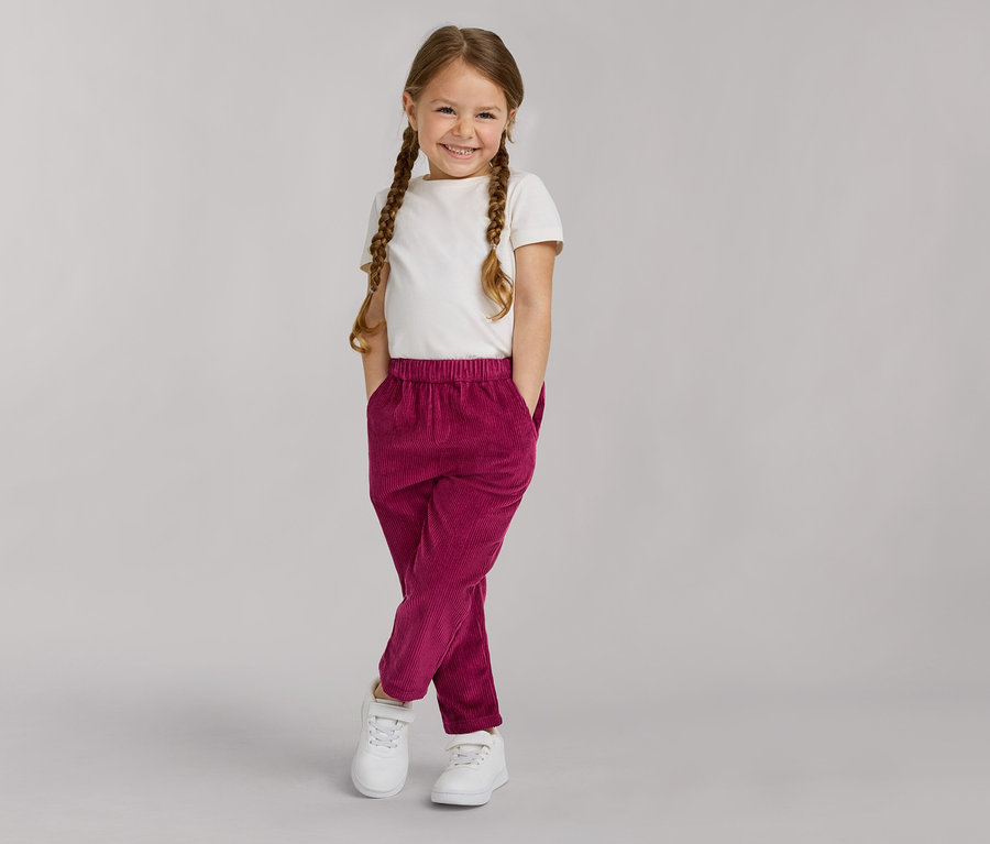 Ein junges Mädchen mit geflochtenen Zöpfen lächelt, trägt ein weißes T-Shirt, magentafarbene Cordhose und weiße Turnschuhe, Hände in den Taschen.