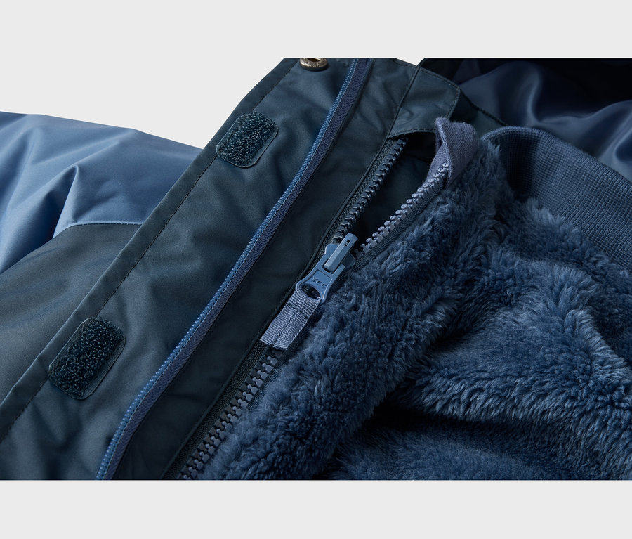 Nahaufnahme einer blauen Jacke mit dunklem Oberstoff, teilweise geöffnetem Reißverschluss und flauschigem Fleecefutter. Klettverschlüsse sind an der Reißverschlussleiste sichtbar.