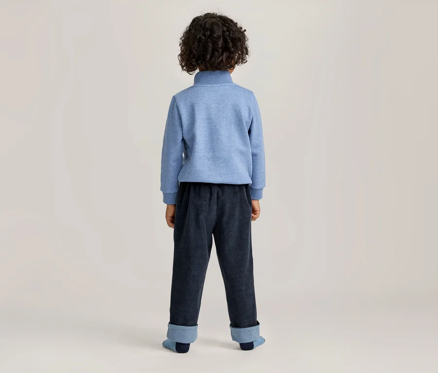 Kind von hinten, trägt einen blauen Pullover mit Stehkragen und dunkelblaue Cordhose mit umgeschlagenen, hellblauen Bündchen.