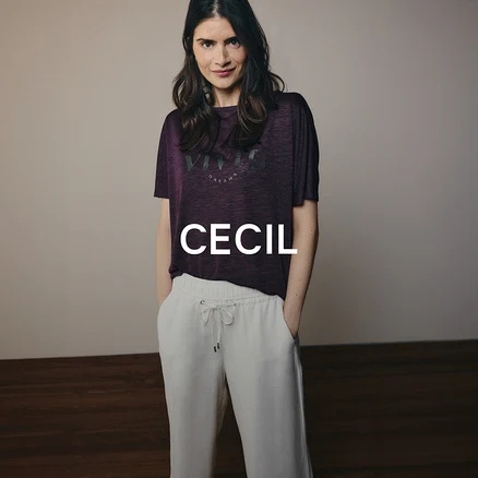 Eine Frau mit dunklen Haaren trägt ein lila T-Shirt mit Aufschrift und eine helle Hose mit Kordelzug. Sie steht mit den Händen in den Hosentaschen und blickt in die Kamera.