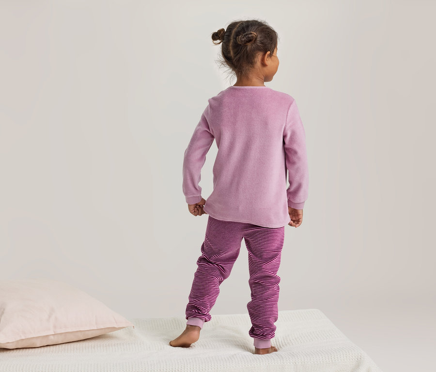 Kind von hinten, in rosa Pyjama mit gestreiften Hosen, steht auf einer weißen Decke neben einem hellrosa Kissen.