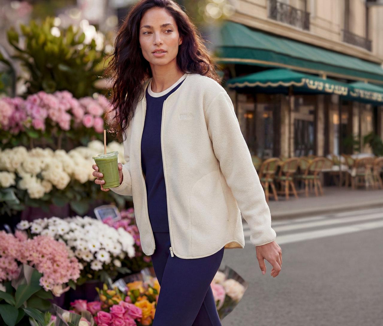 Frau geht an Blumenstand und Café vorbei, hält einen grünen Smoothie. Sie trägt eine cremefarbene Fila-Fleecejacke, ein marineblaues Oberteil und Leggings.