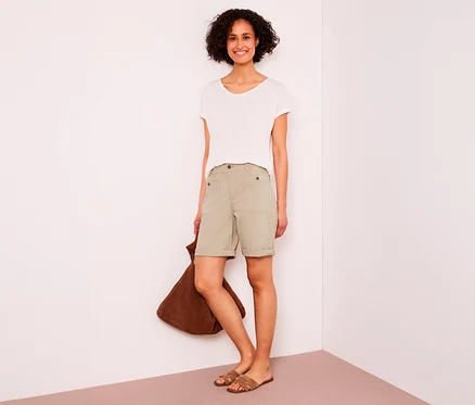 Lachende Frau mit kurzen Locken trägt ein weißes T-Shirt, beigefarbene Street One Bermuda „Yulius“ und braune Sandalen, eine braune Tasche haltend.
