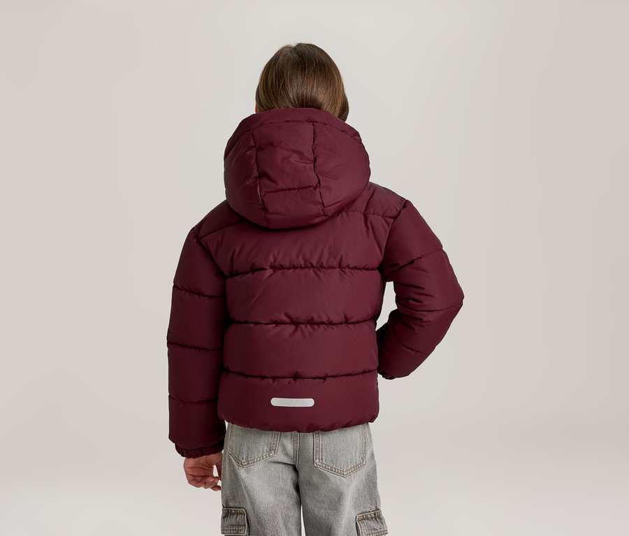 Kind von hinten in einer bordeauxroten Steppjacke mit Kapuze und hellgrauen Cargohosen. Auf dem Rücken der Jacke befindet sich ein reflektierender Streifen.