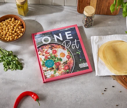 Kochbuch 'One Pot' liegt auf einer Küchentheke, umgeben von Kichererbsen, Koriander, Chili und Tortillas.