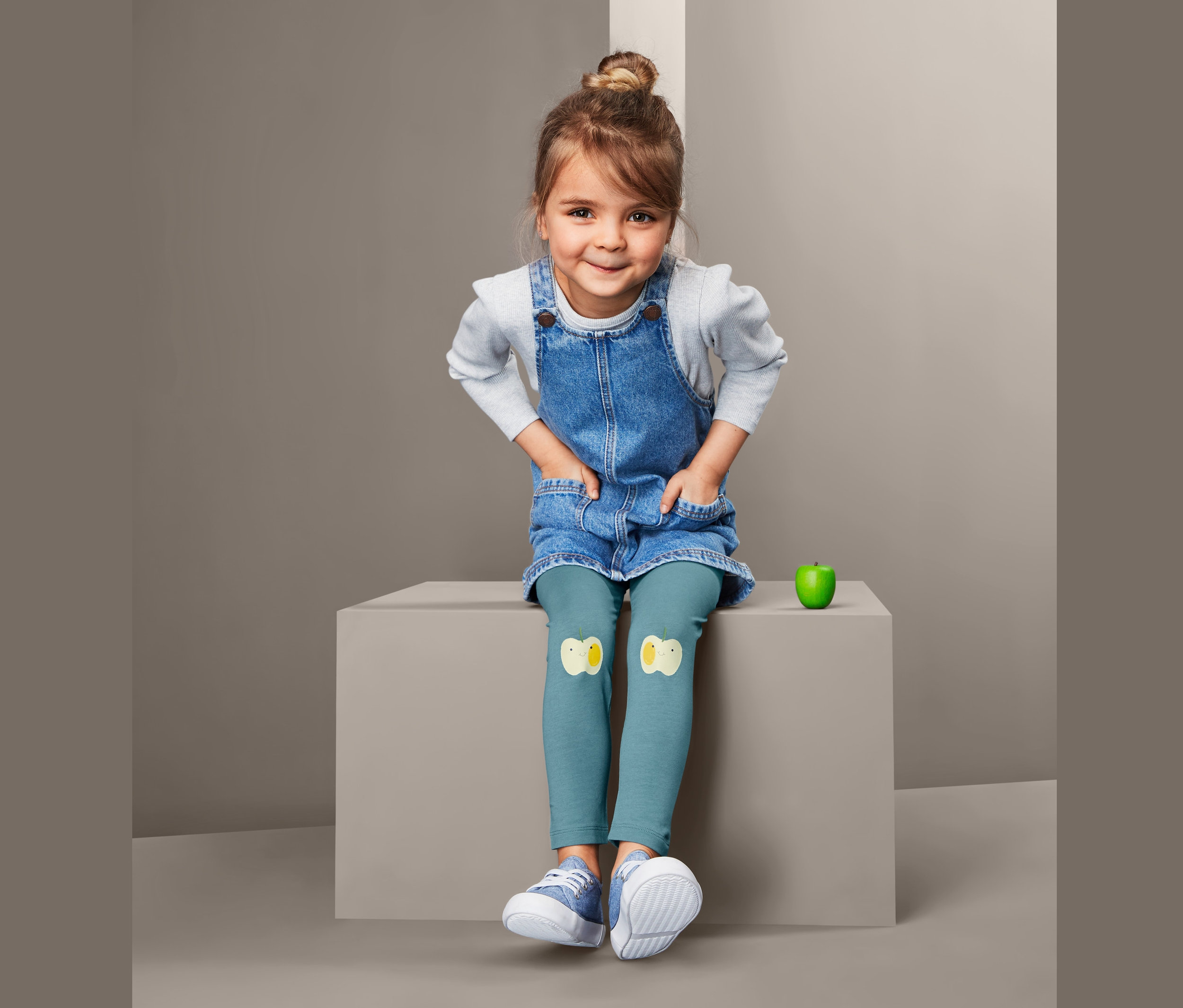 Ein Mädchen sitzt und trägt ein Jeanskleid und 2 Leggings mit Küken-Applikationen.