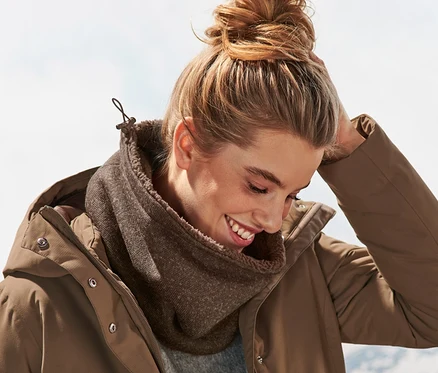 Eine lächelnde Frau mit Dutt trägt einen braunen Strickfleece-Schlauchschal und eine braune Jacke. Im Hintergrund sind Berge zu sehen.