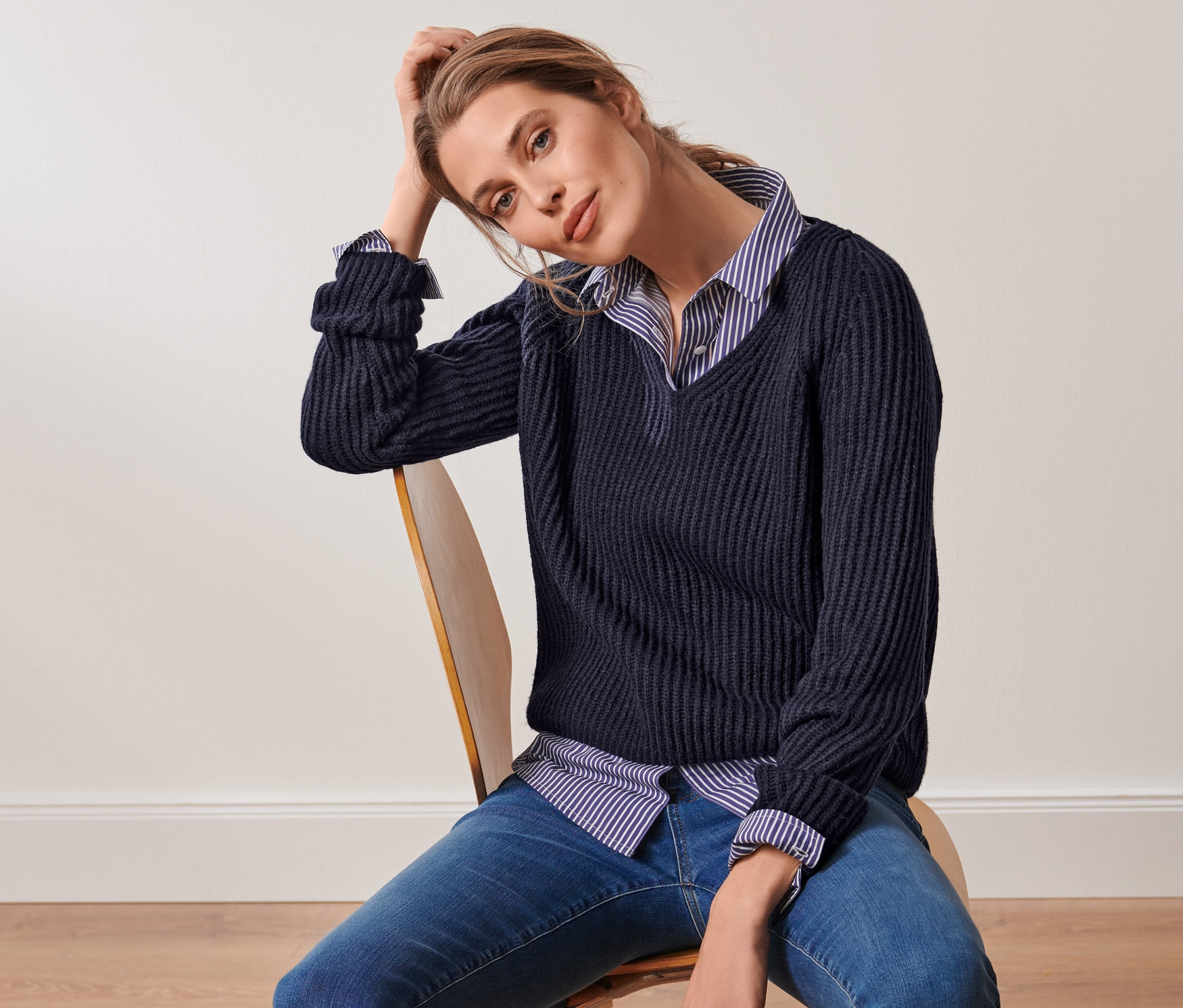 Eine Frau sitzt auf einem Stuhl und trägt eine gestreifte Hemdbluse und einen dunkelblauen Grobstrick-Pullover mit Wolle.