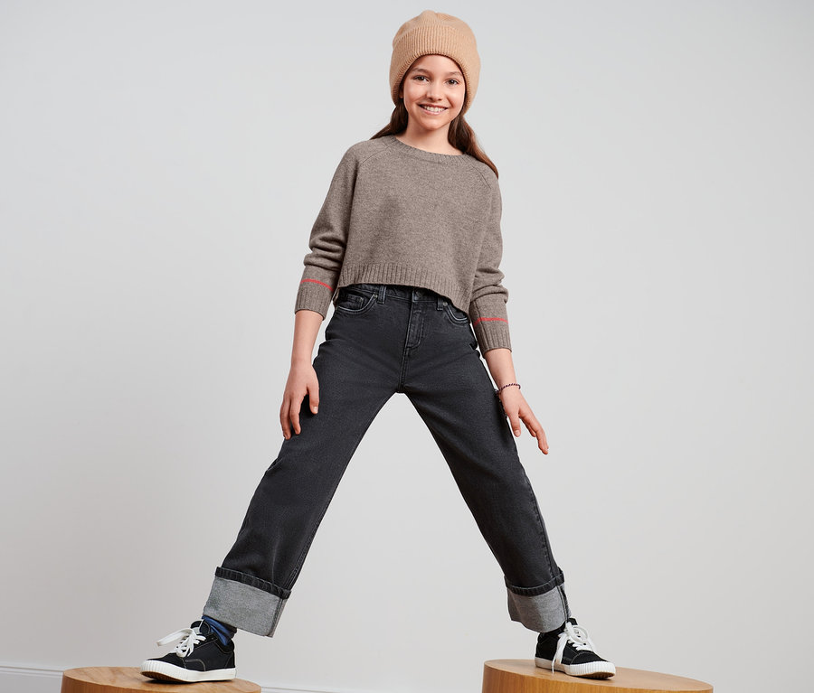 Lächelndes junges Mädchen steht auf Holzhockern und trägt eine Bootcut-Jeans, einen braunen Pullover und eine braune Mütze.