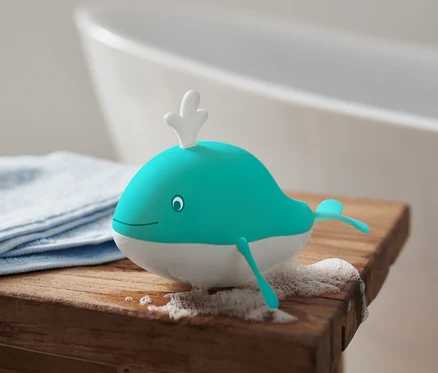 Badespielzeug Aufziehwal liegt auf einem Holzhocker neben einem blauen Tuch und Schaum, im Hintergrund eine Badewanne.