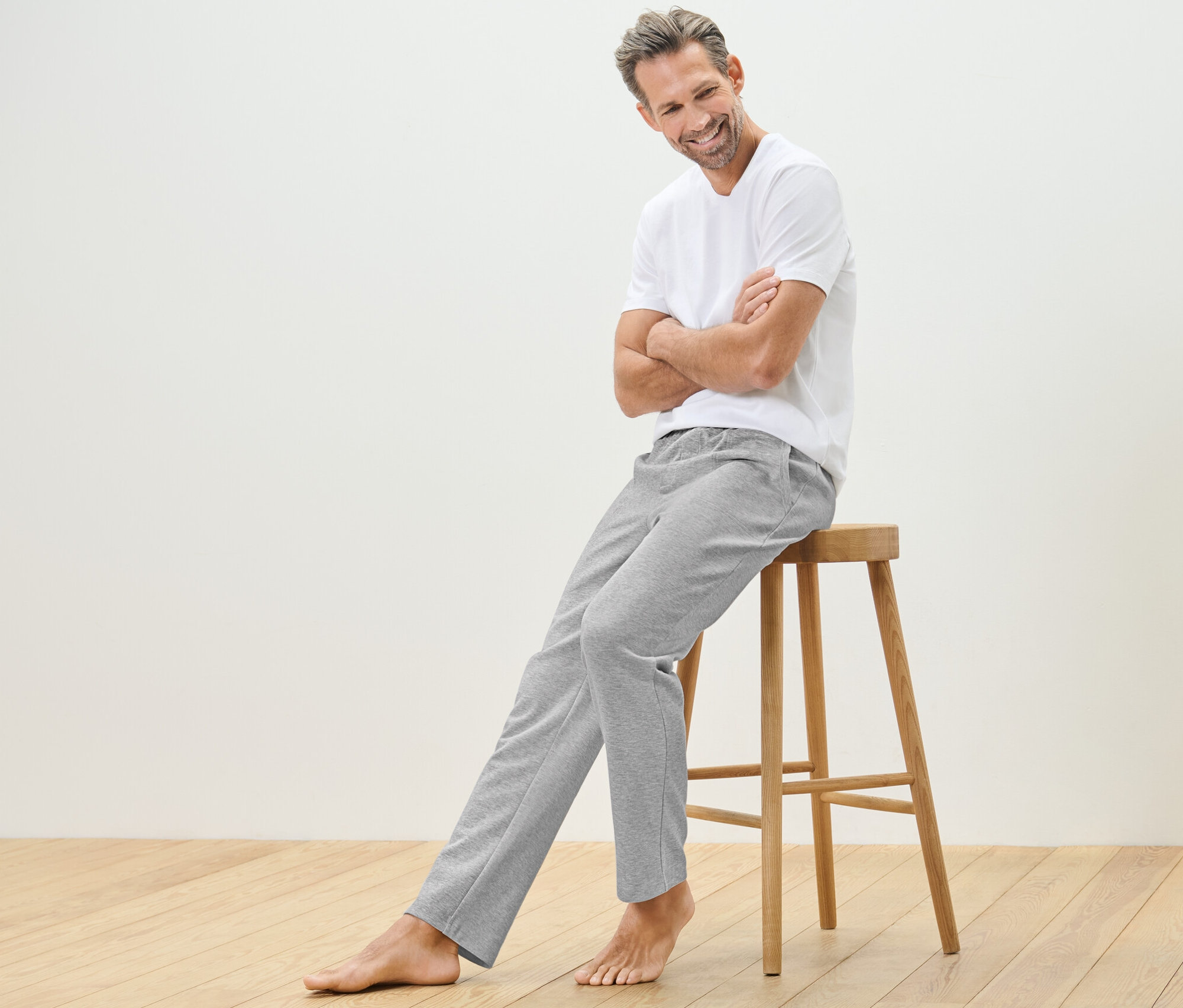 Mann sitzt auf einem Holzhocker und trägt eine Homewearhose und ein T-Shirt.