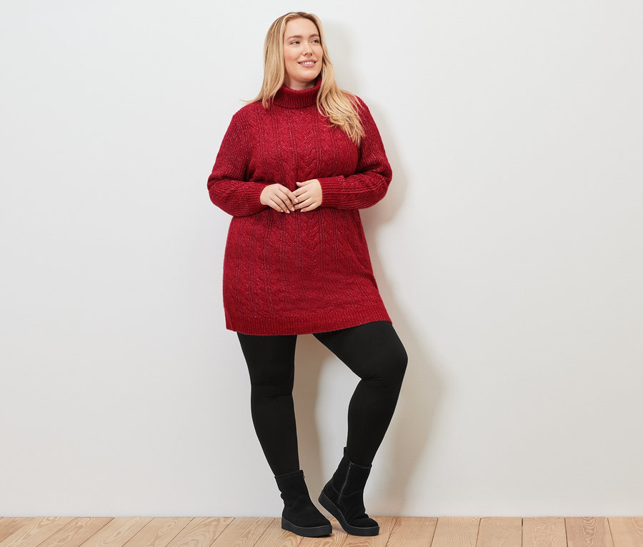 Frau posiert in langem, rotem Strickpullover mit Zopfmuster, schwarzen Leggings und Stiefeln.