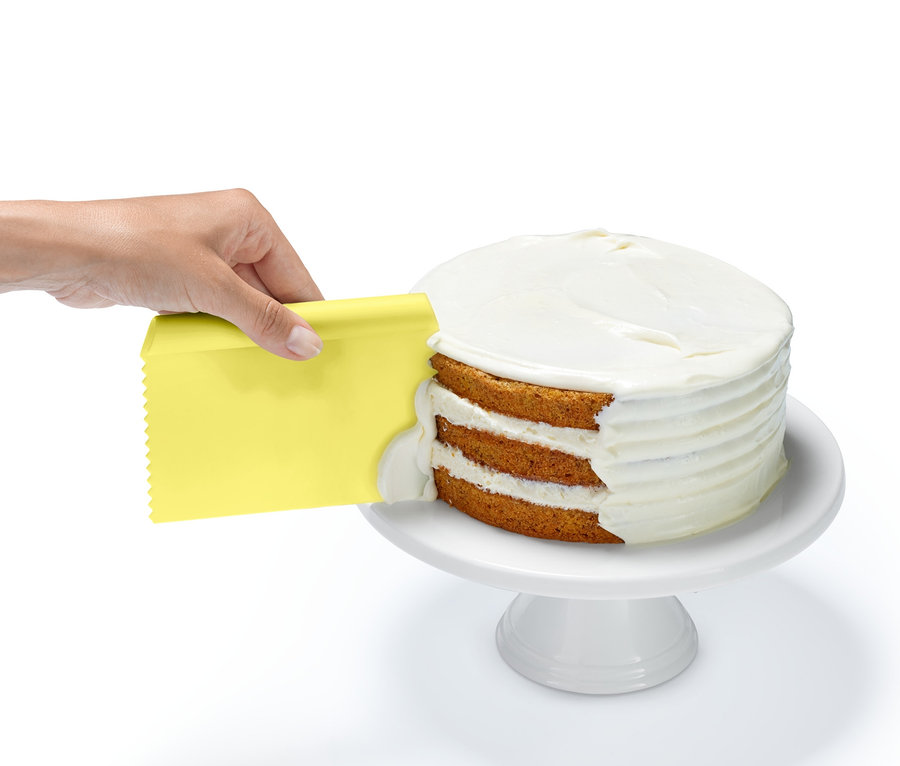 Eine Hand verwendet einen gelben Schaber aus dem Backhelfer-Set, um eine weiße Glasur auf die Schichten eines Kuchens aufzutragen.