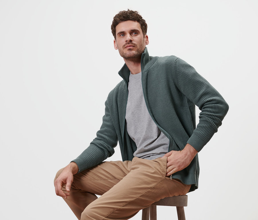 Mann sitzt auf einem Hocker und trägt eine Strickjacke in Salbei.