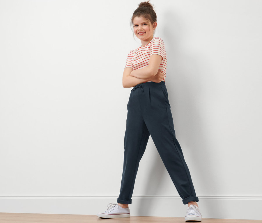 Lächelndes Mädchen posiert in Kinder-Sweathose, rot gestreiftem T-Shirt und weißen Schuhen.
