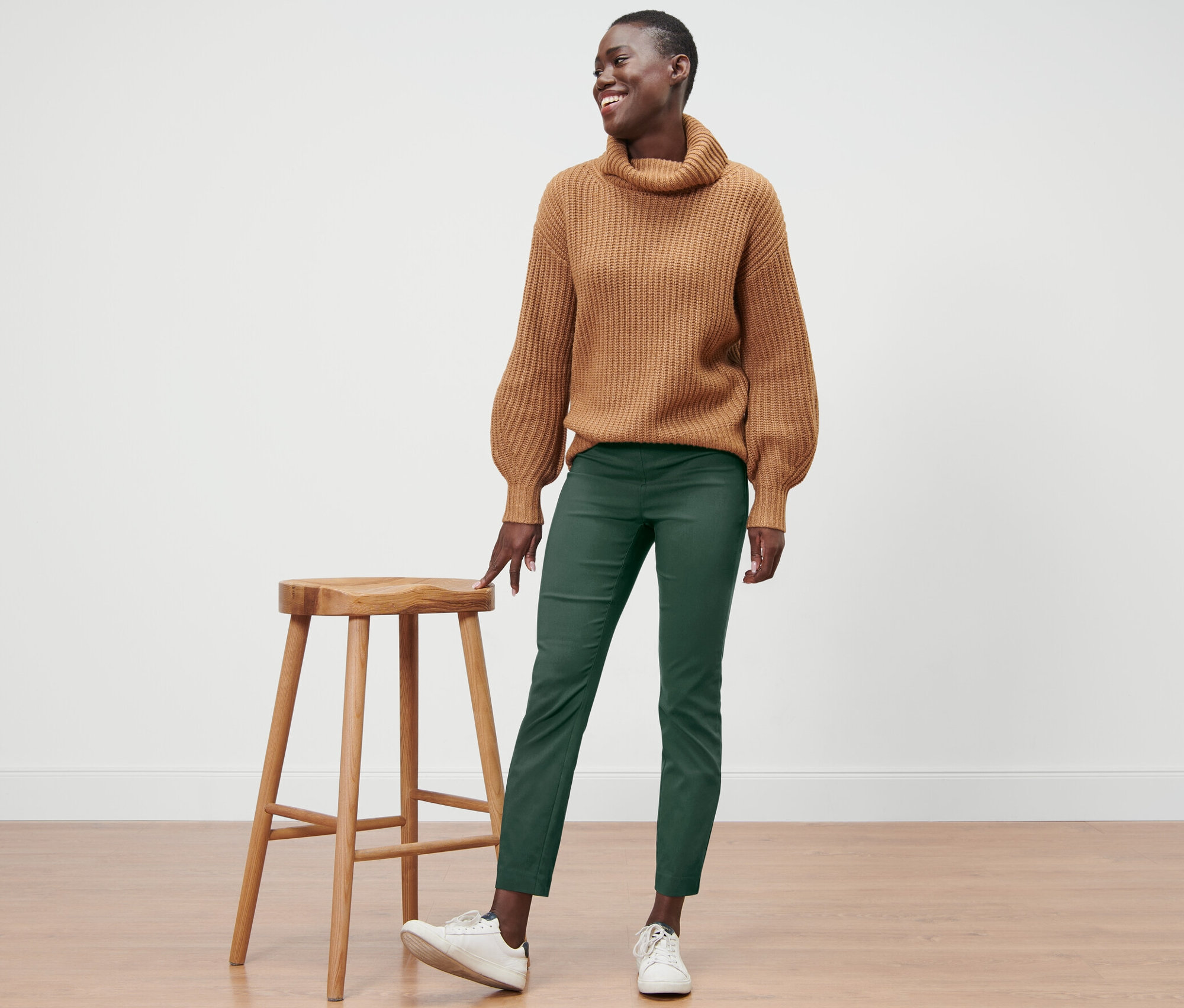 Eine lächelnde Frau steht neben einem Holzhocker und trägt einen braunen Pullover und eine grüne Stretchhose.