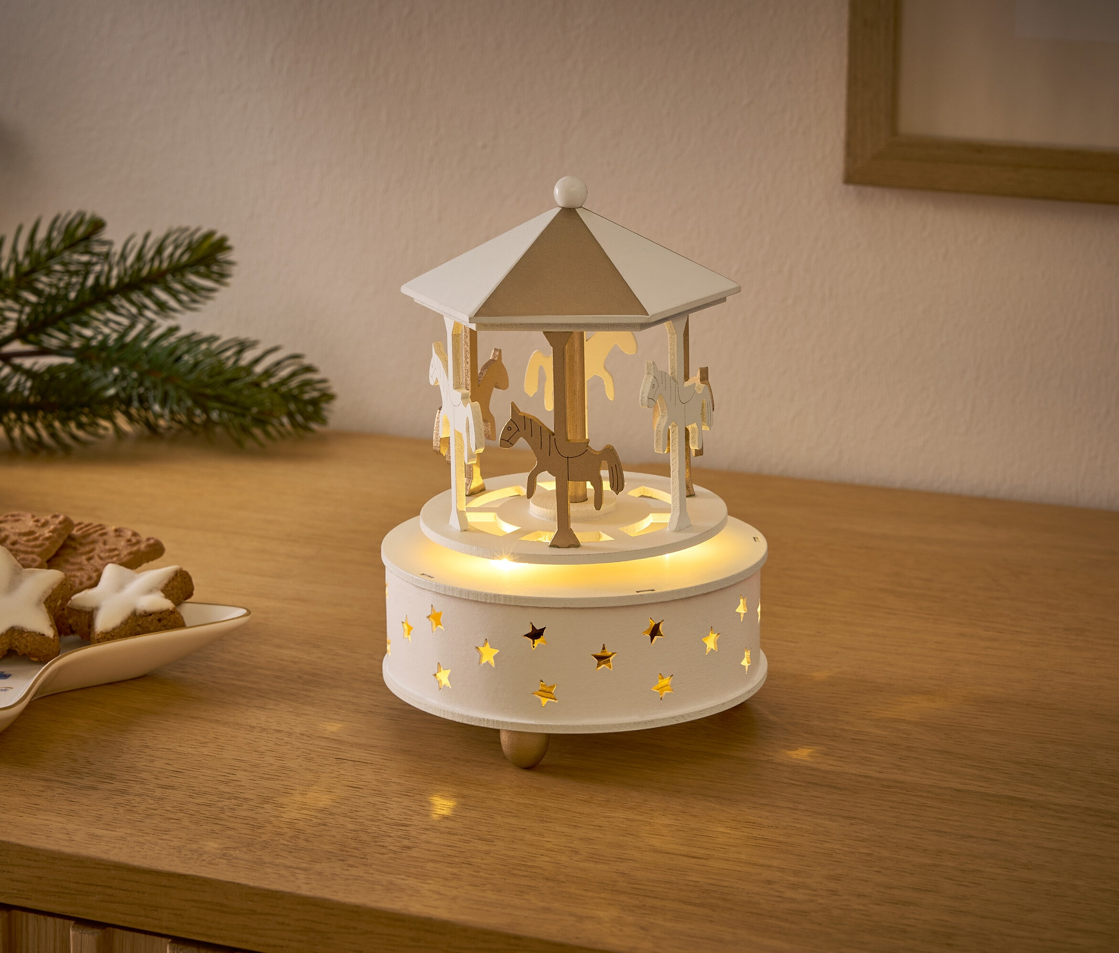 Ein hölzernes Karussell mit Pferden und Sternen leuchtet auf einem Holztisch neben Weihnachtsplätzchen.