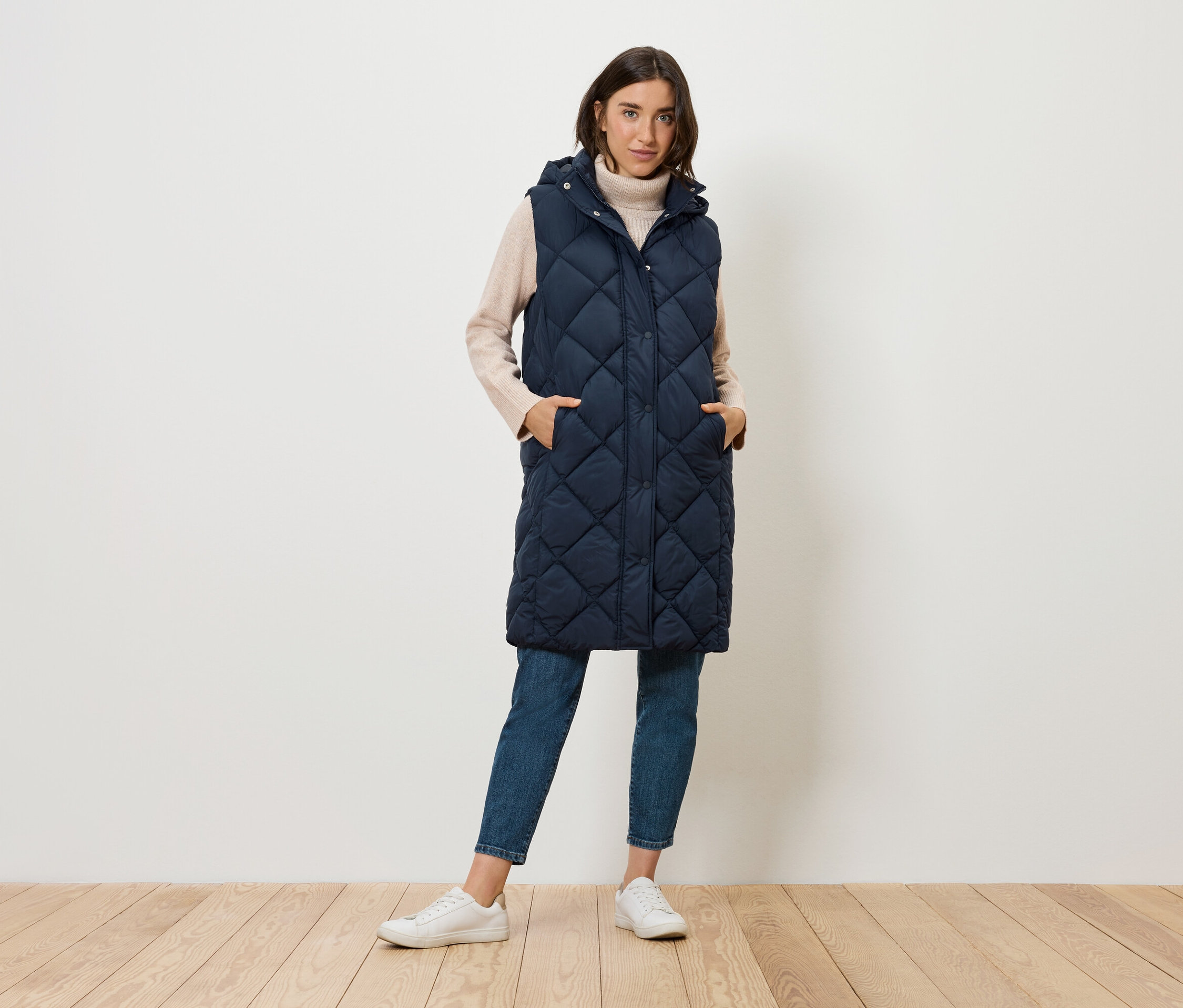 Eine Frau steht und trägt eine lange, gesteppte, marineblaue Weste mit Kapuze, Jeans und Turnschuhe.