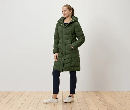 Eine Frau in einer langen, grünen Steppjacke mit Kapuze und dunkelblauen Hosen steht auf einem Holzboden.