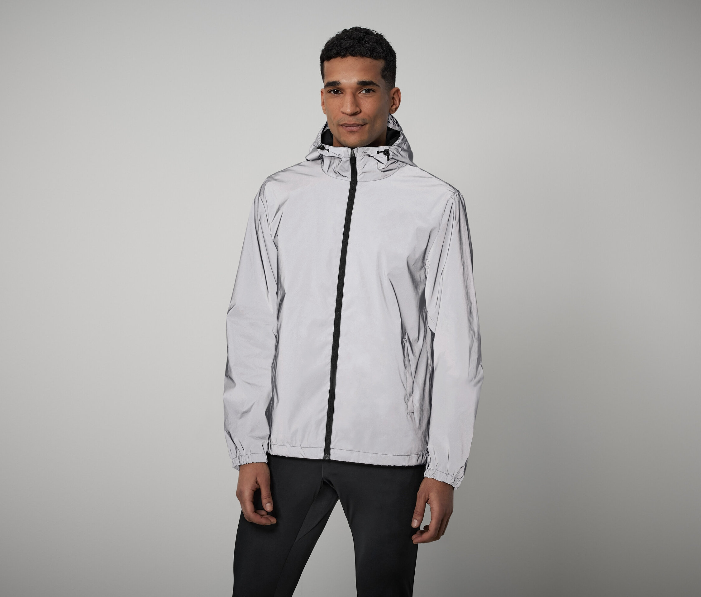 Mann steht und trägt eine graue, reflektierende Reißverschlussjacke mit Kapuze und eine schwarze Hose.
