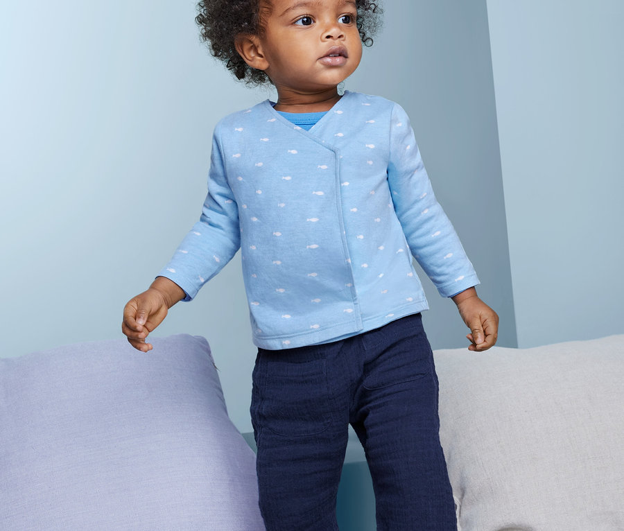 Ein dunkelhäutiges Kleinkind steht. Es trägt eine blaue 2 Baby Baumwoll-Wickeljacke.