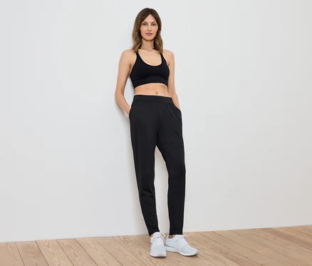 Eine Frau in einem schwarzen Sporttop und Hosen steht an einer weißen Wand.