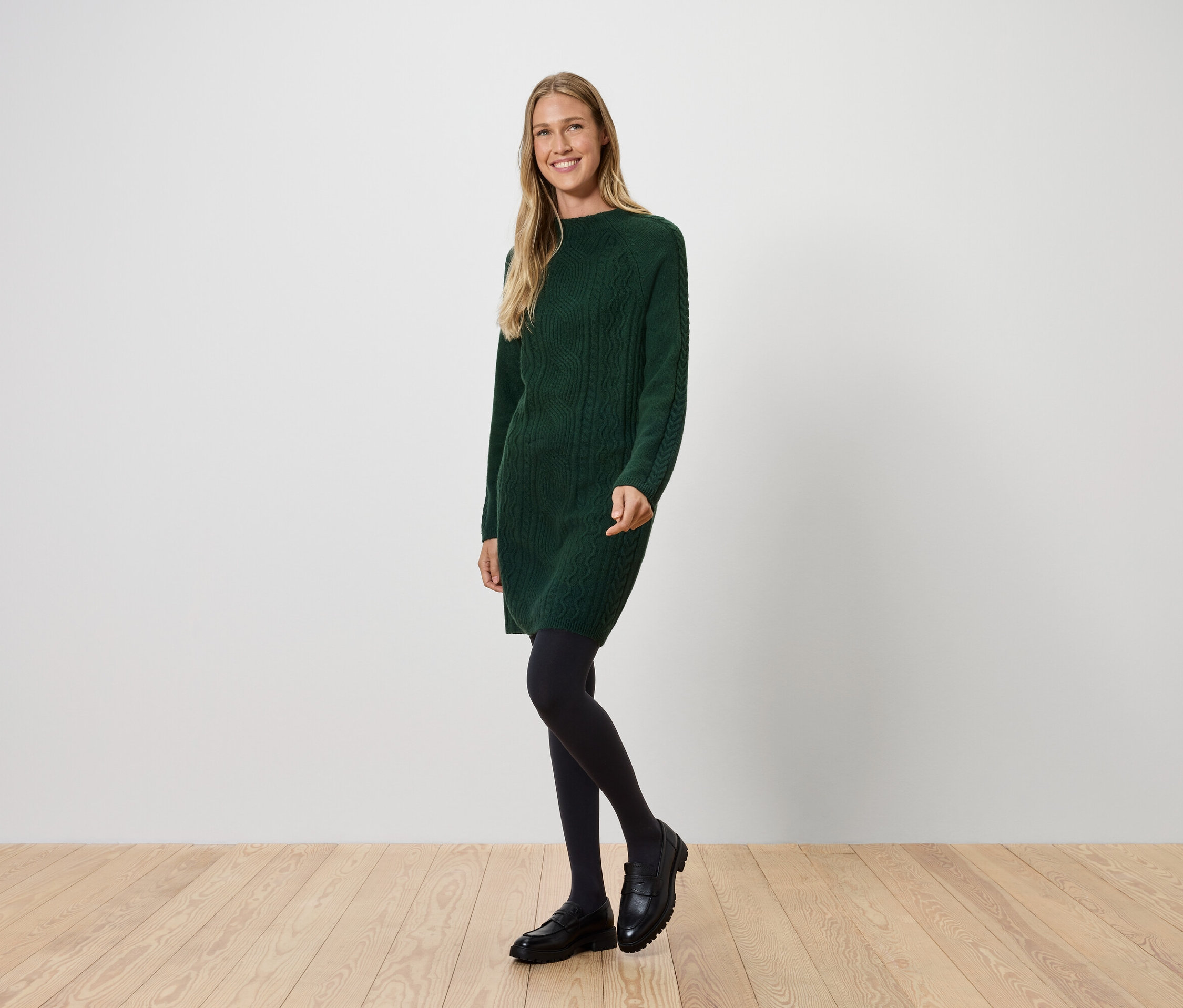 Frau in grünem Strickkleid, schwarzer Strumpfhose und schwarzen Schuhen posiert auf Holzboden vor einer weißen Wand.