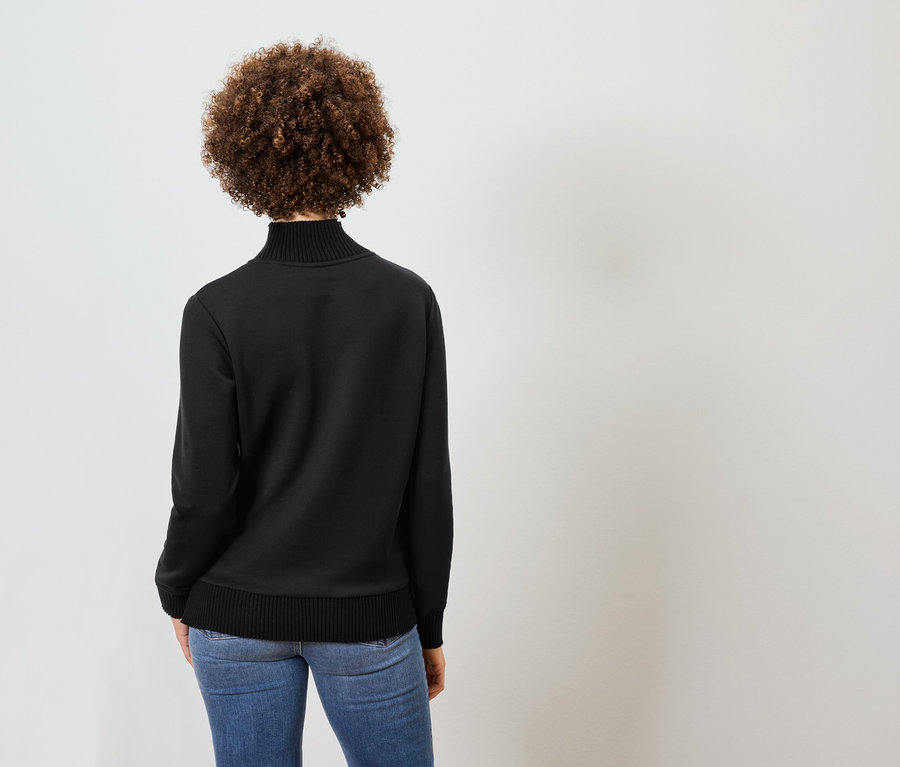 Rückansicht einer Frau mit lockigem Haar, die einen schwarzen Rollkragenpullover und blaue Jeans trägt.
