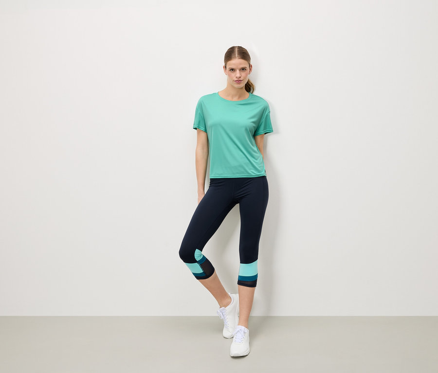 Frau steht vor einer weißen Wand und trägt ein türkisfarbenes T-Shirt, marineblaue Leggings mit türkisfarbenen Details und weiße Turnschuhe.